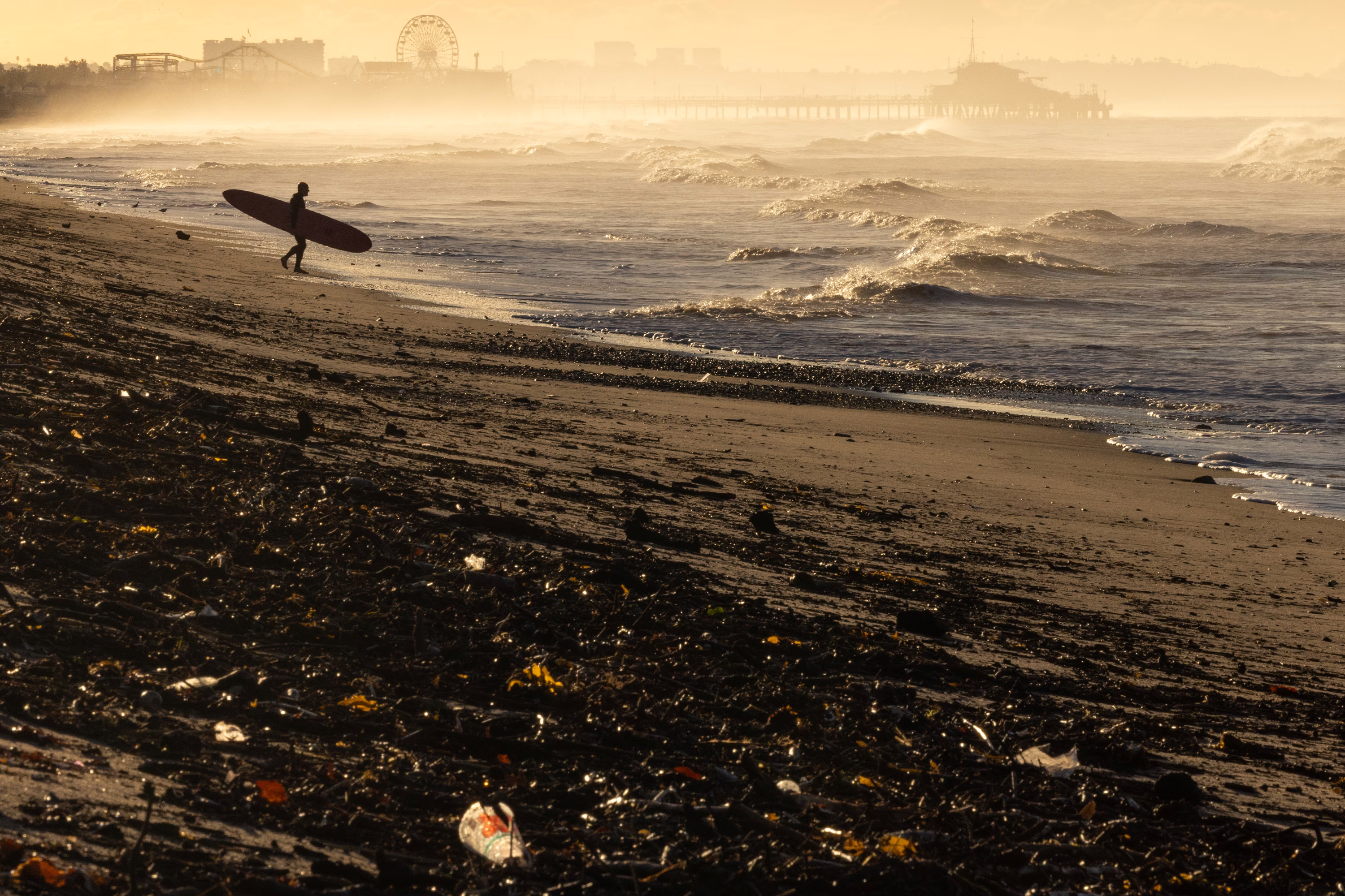 A person carries a surfboard toward the shoreline, walking on a beach that is partly covered with debris from a previous wildfire