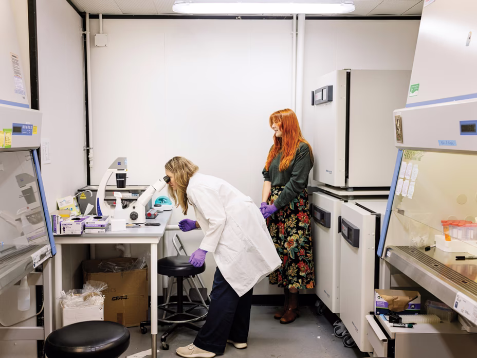 author wearing white coat in laboratory bends to look into an instrument with another scientist standing and supervising in background