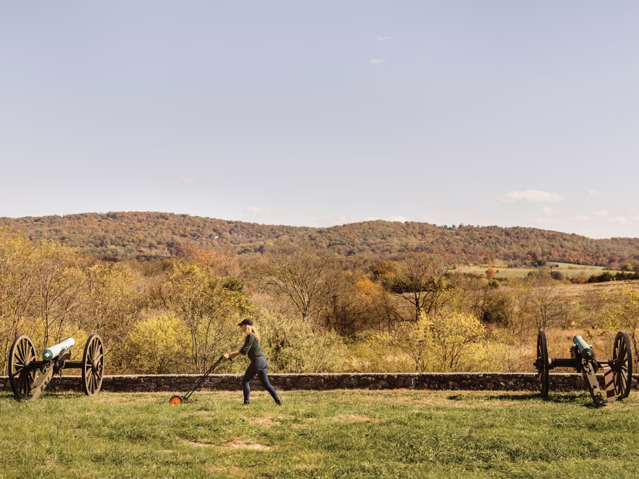 photo of the author using hand mower to mow grass between two Civil-War-era cannons on battlefield with hill in distance
