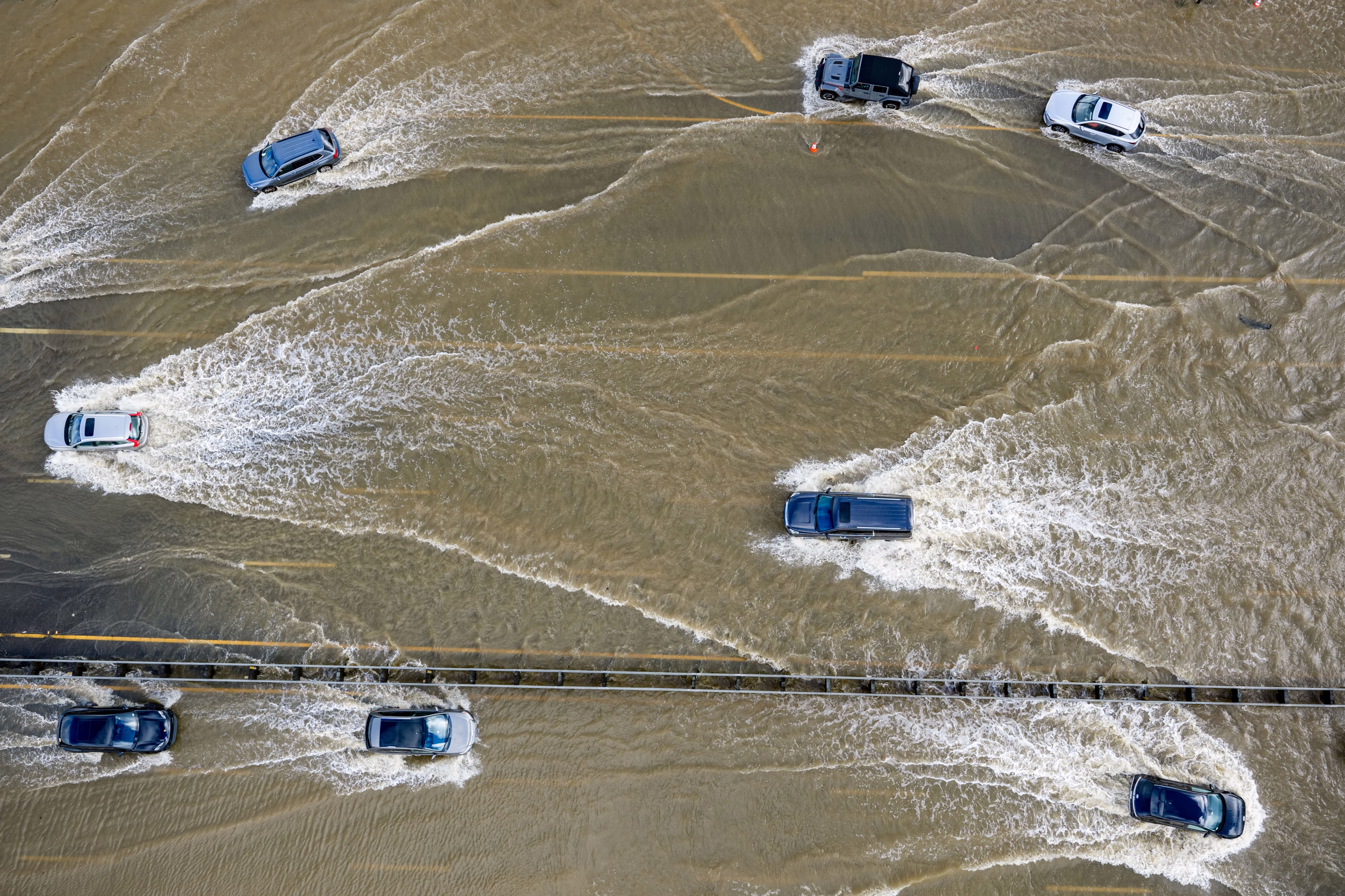 An aerial view of several cars driving on a highway that has covered by shallow floodwater, each car leaving a wake behind it.