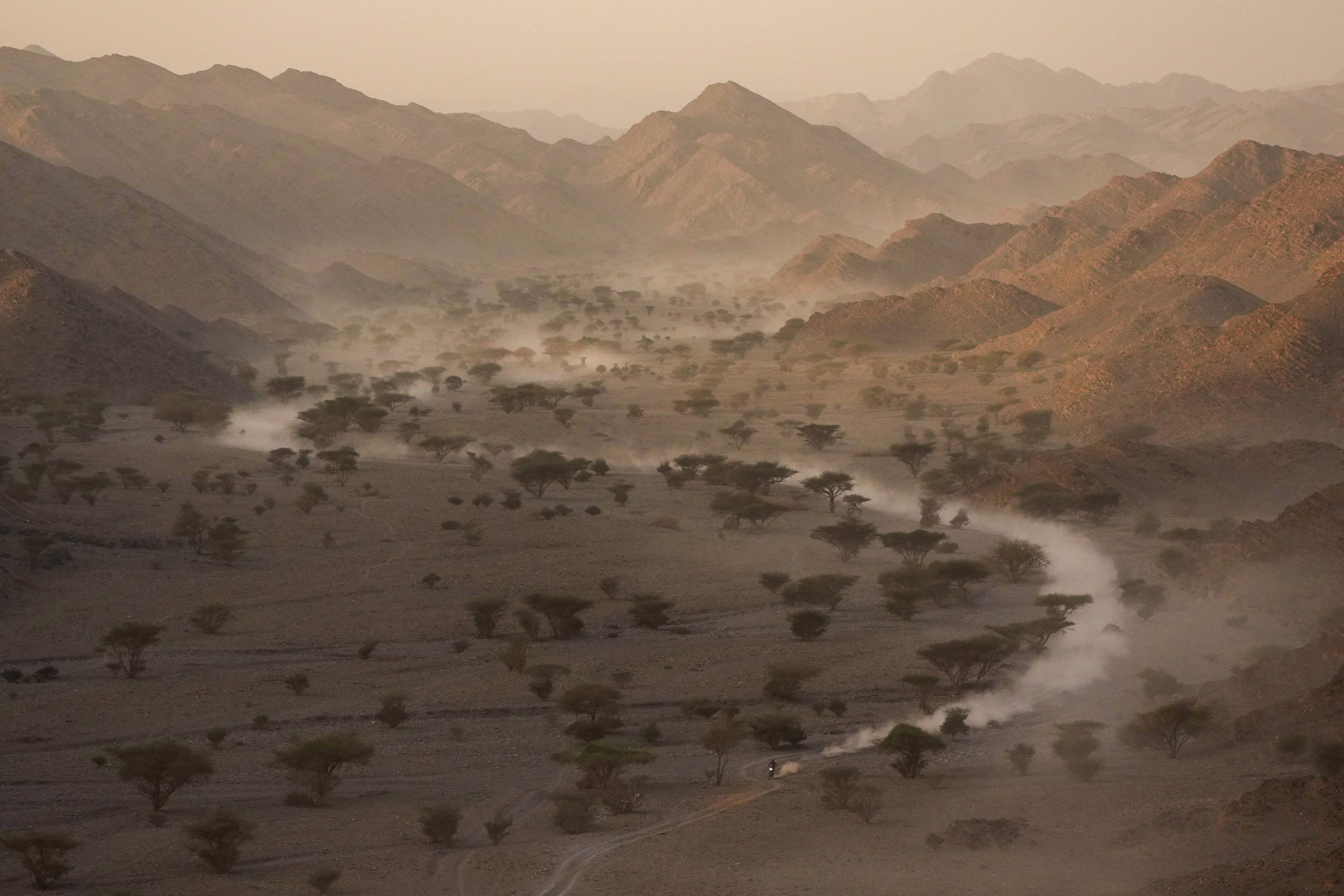 A motorcycle racer rides on a dusty path through a shallow desert valley.