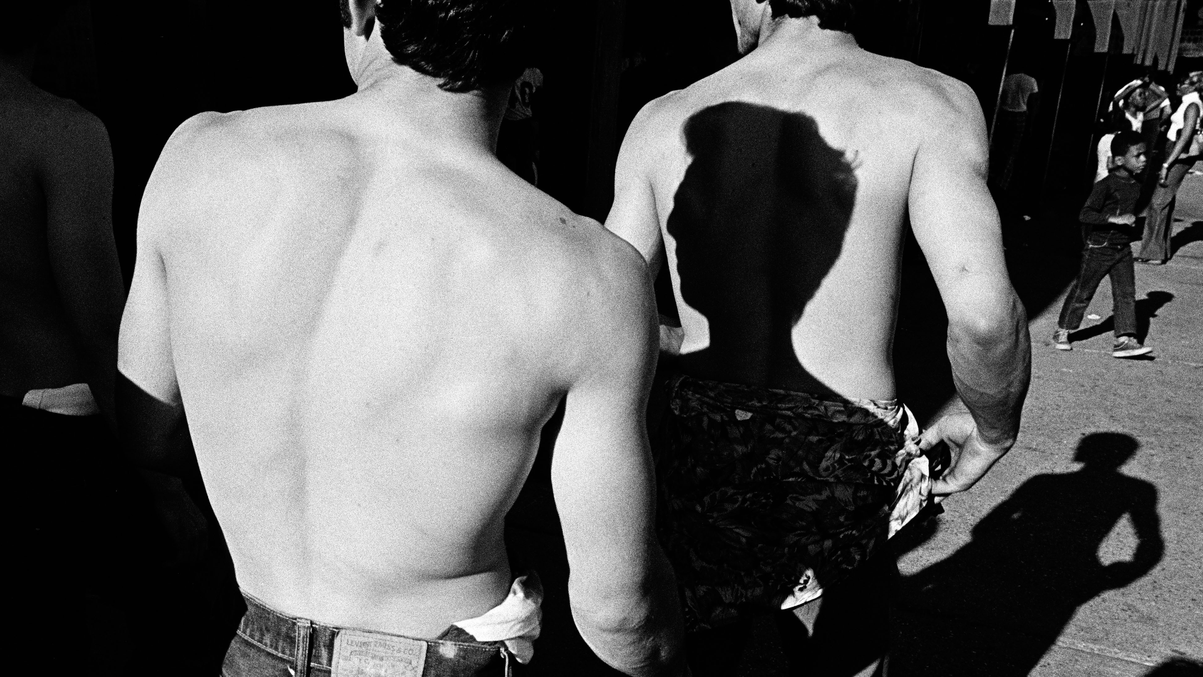 Black-and-white photo of one muscle-bound man standing behind another, casting a shadow on his back.