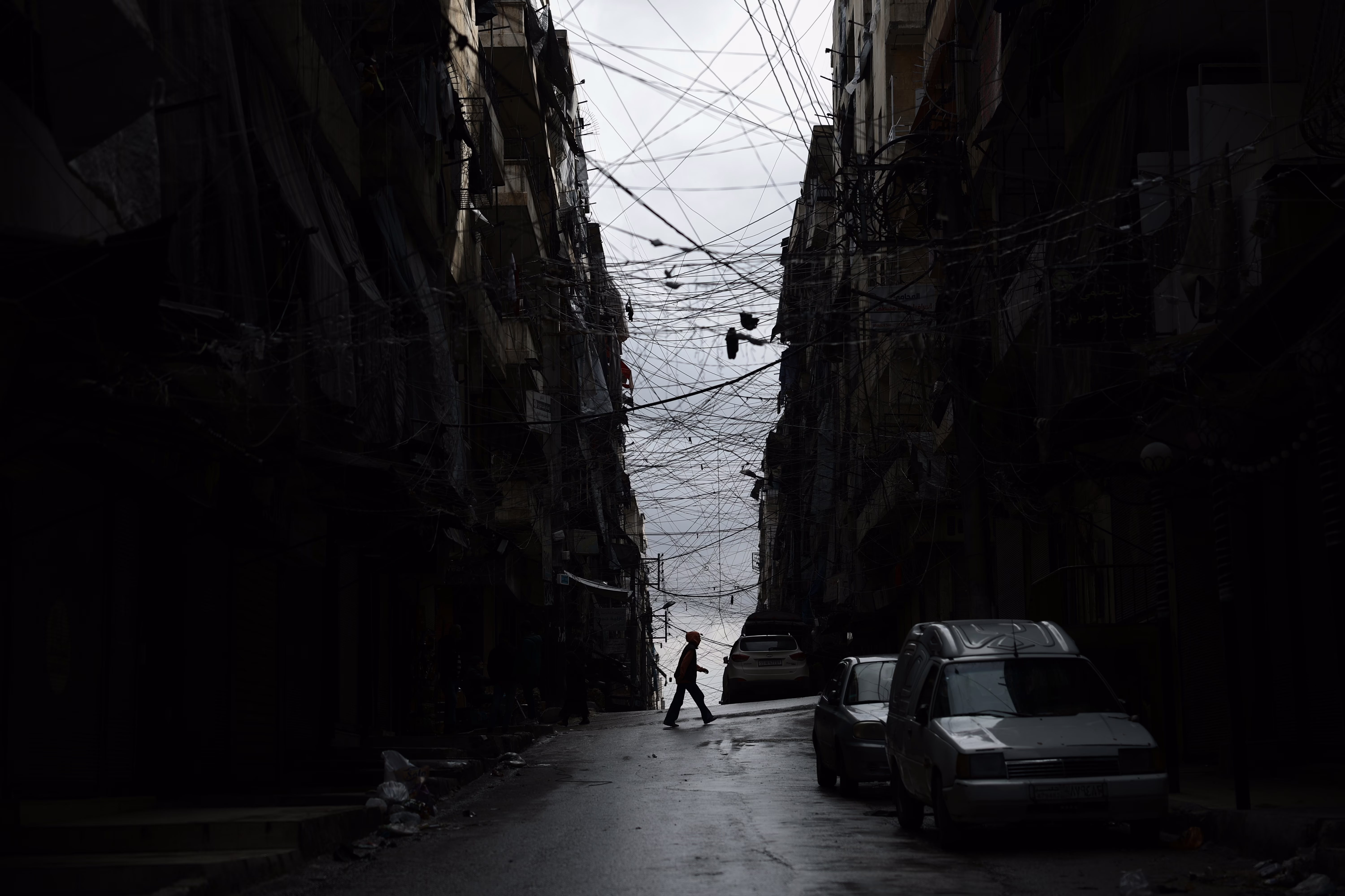 A man crosses a street, flanked by tall, dark buildings and a chaotic web of wires above.
