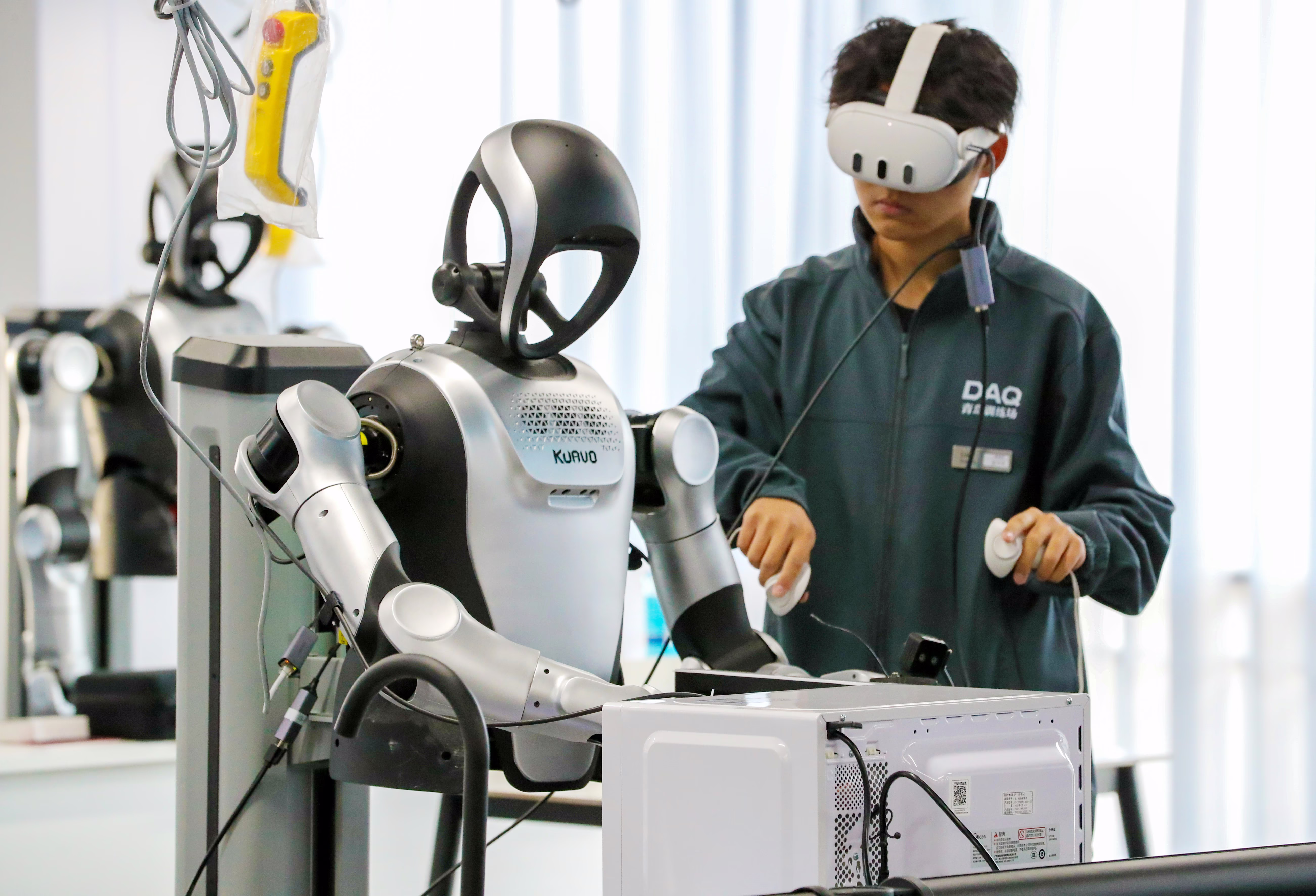 A person wearing VR goggles stands beside a humanoid robot, training it to clean a microwave oven.