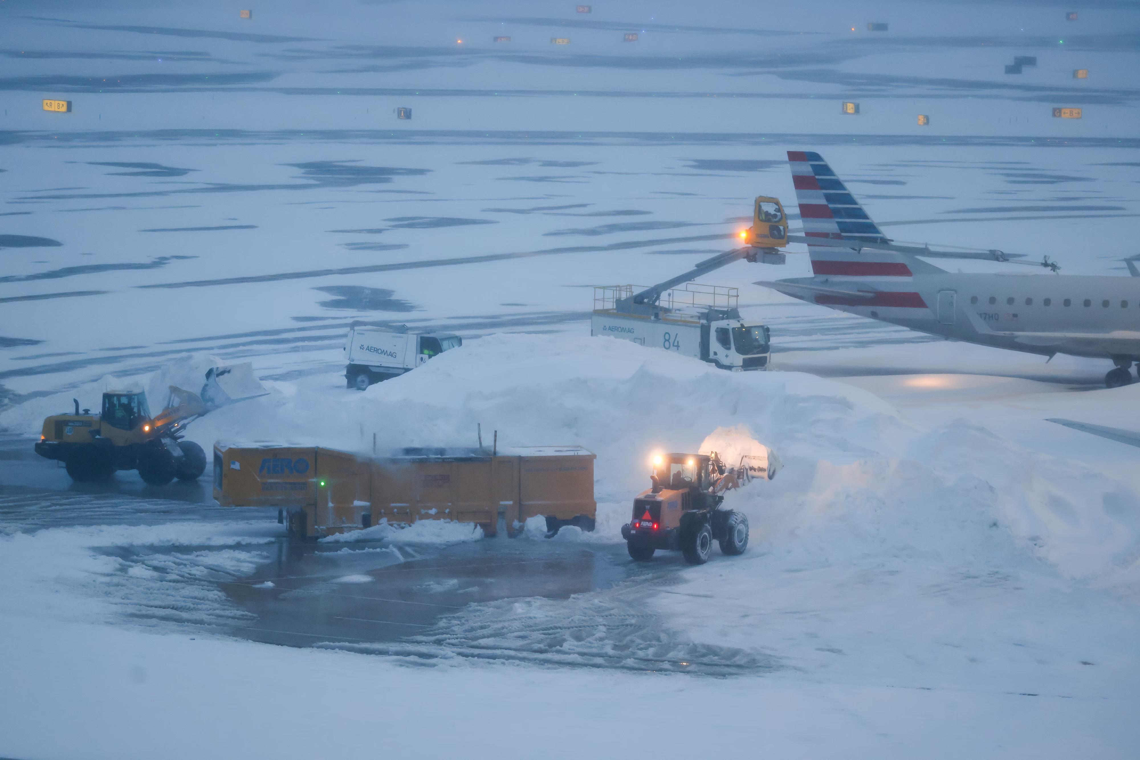 Two front-end loaders dump snow into an ice melter near a passenger aircraft.