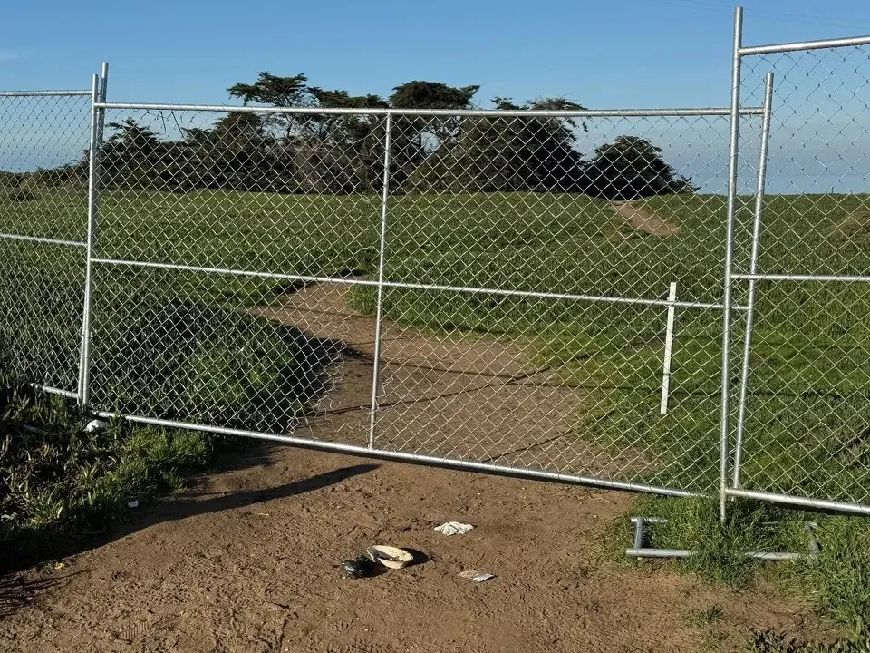 An 8-foot-tall chainlink fence blocks a dirt path leading into a grassy field.
