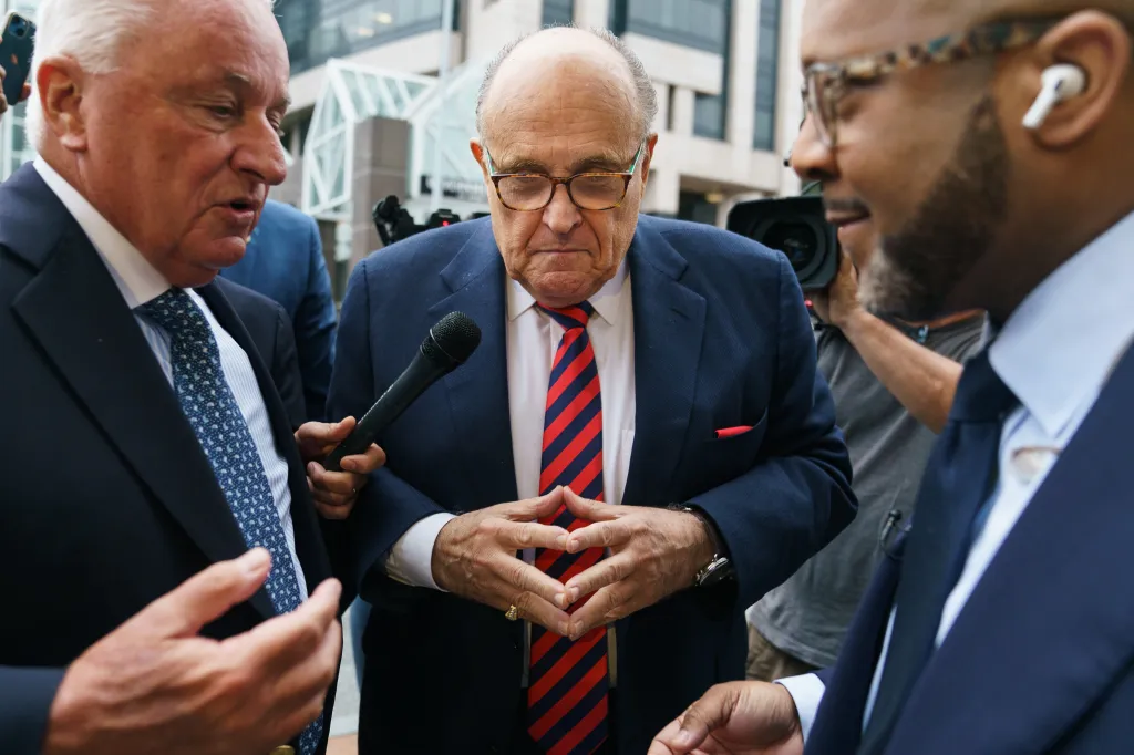 Rudy Giuliani, wearing a red and navy striped tie, stands with his hands clasped as his attorney, Robert Costello, speaks into a microphone.