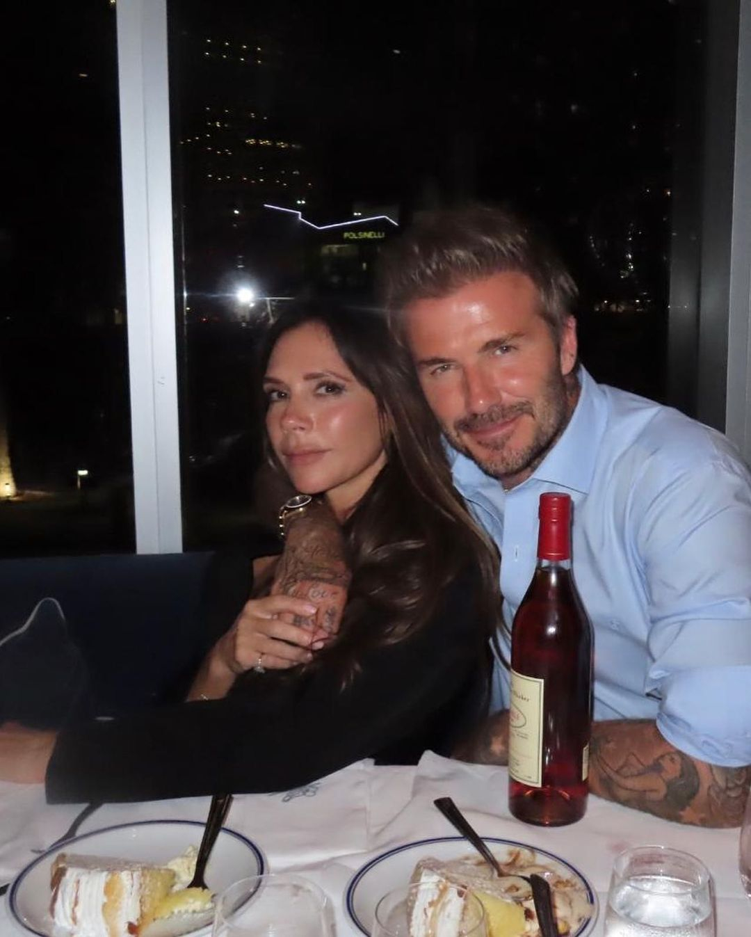 David and Victoria Beckham seated at a table with cake and a bottle of wine.