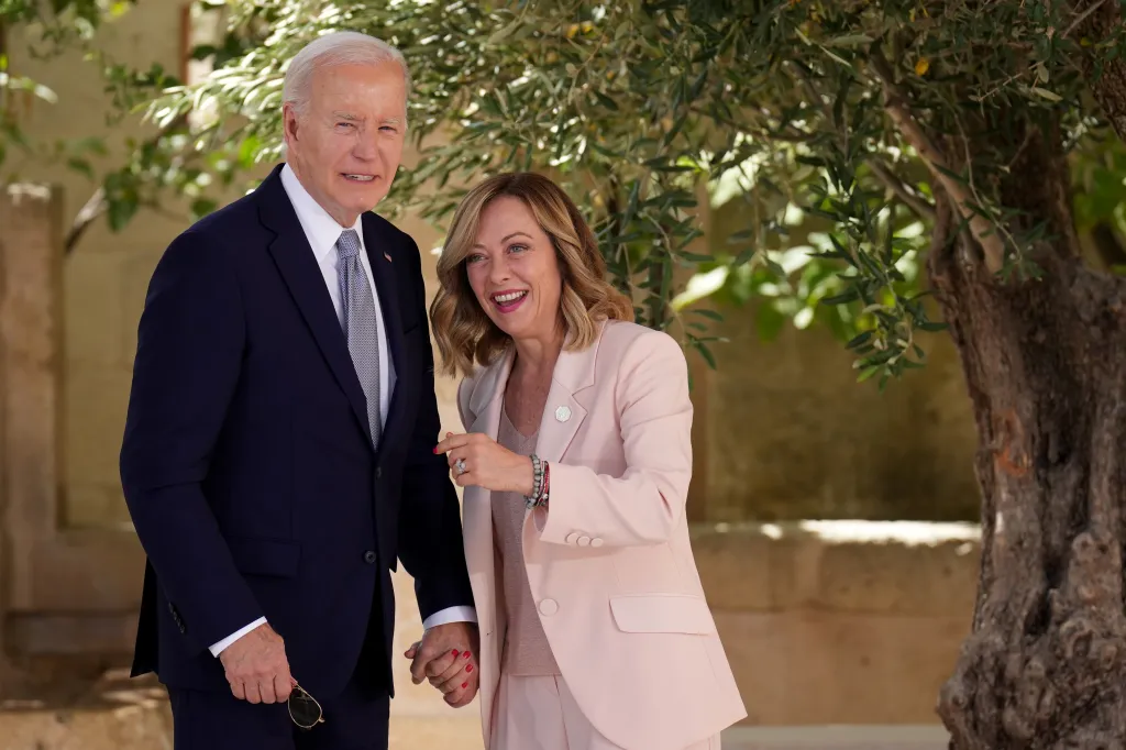 US President Joe Biden and Italian Prime Minister Giorgia Meloni smiling, holding hands, and standing under a leafy tree.