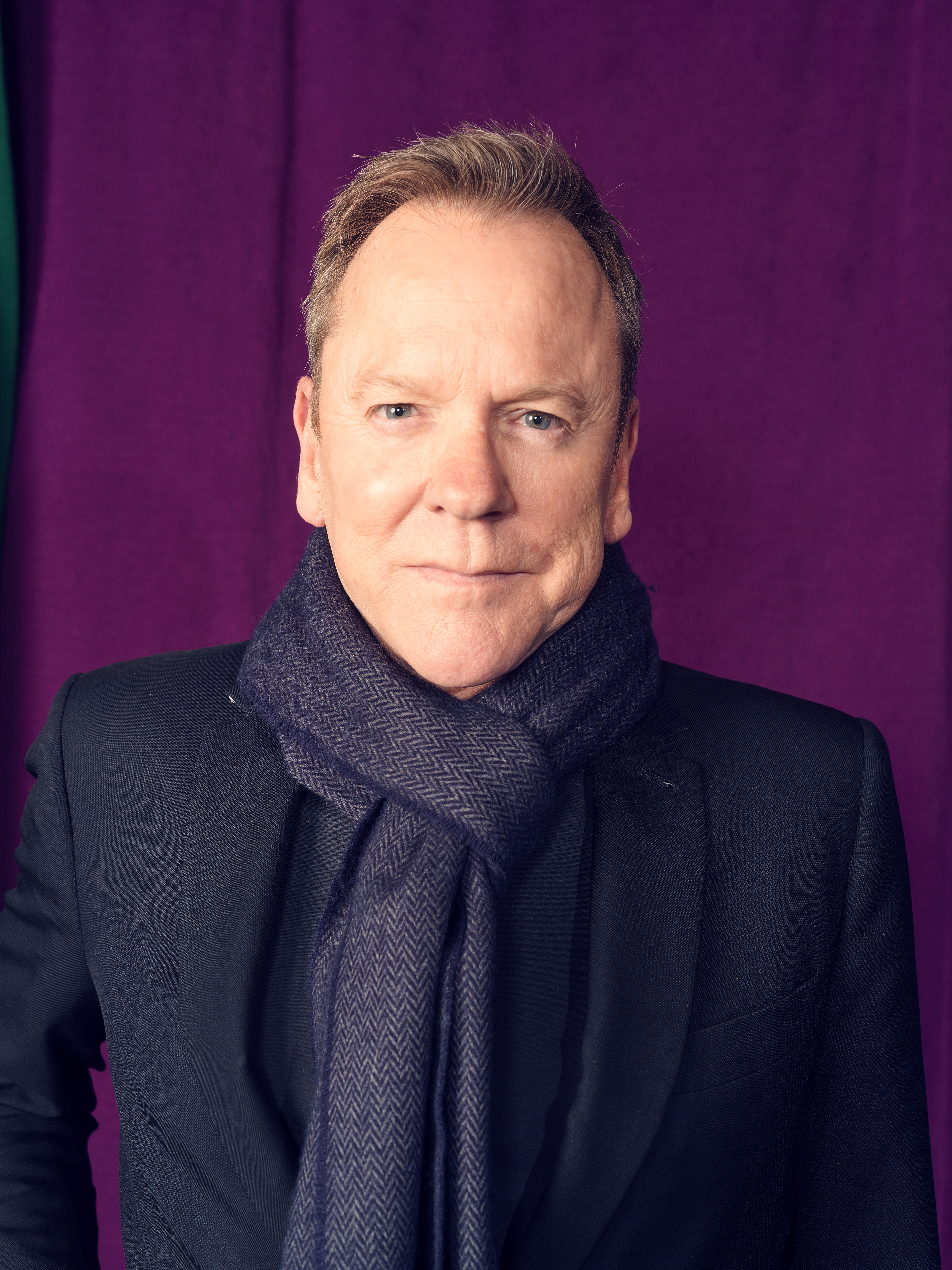 Kiefer Sutherland posing for a portrait against a purple backdrop.