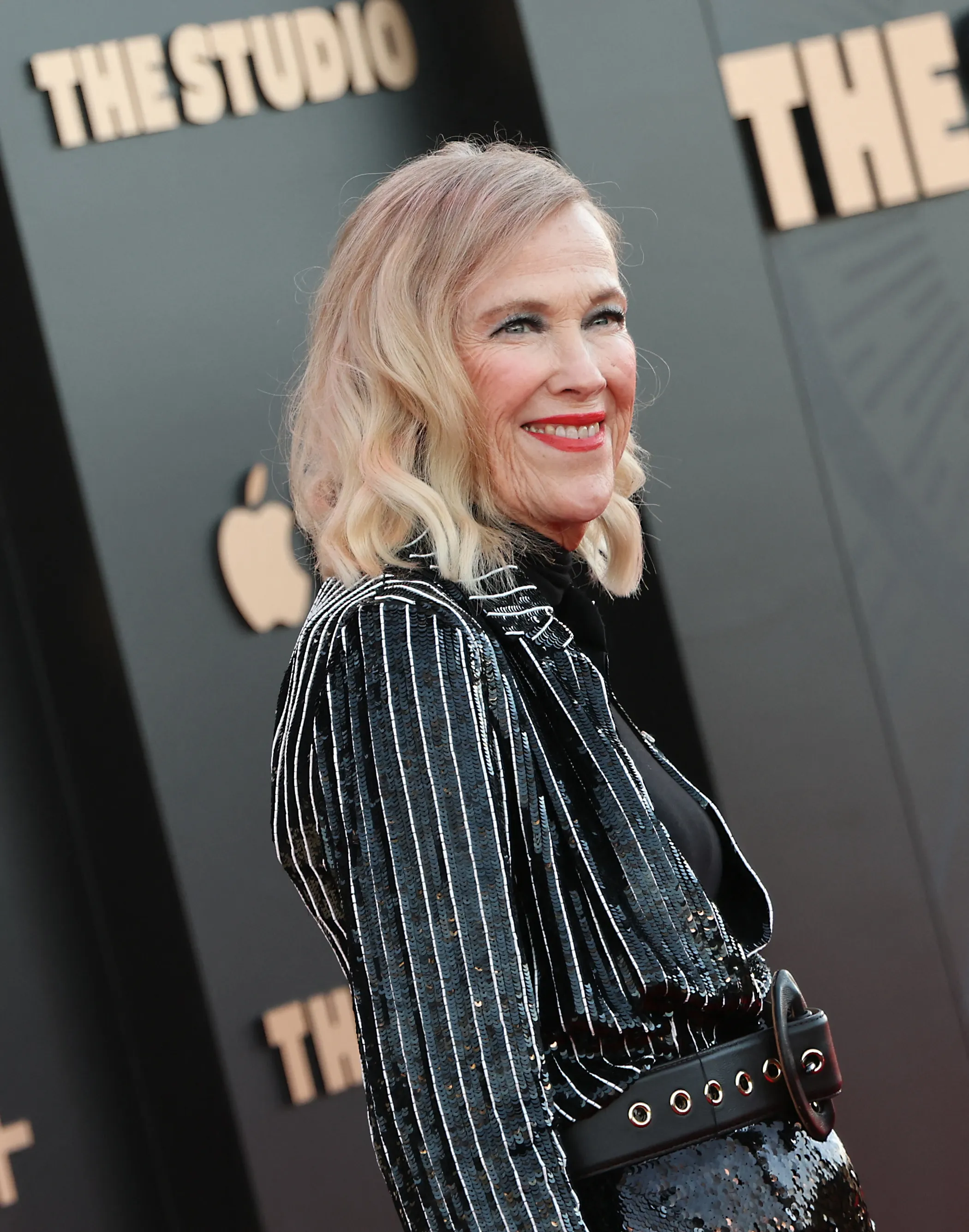 Catherine O'Hara smiles while wearing a sequined jacket with white pinstripes.