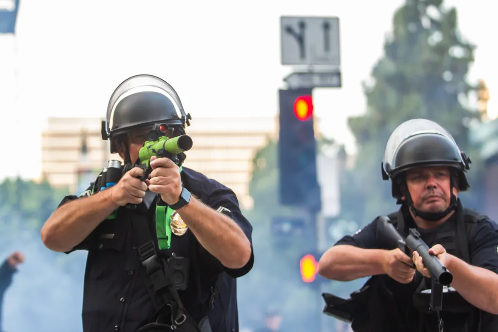 Two Los Angeles Police officers in riot gear point rubber bullet guns at a crowd.