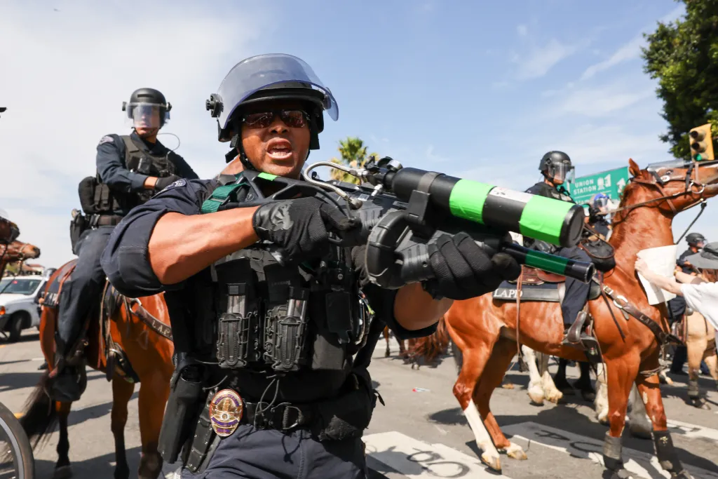 A mounted police officer wearing a helmet and sunglasses holds a riot gun while yelling during an anti-Trump protest in Los Angeles.