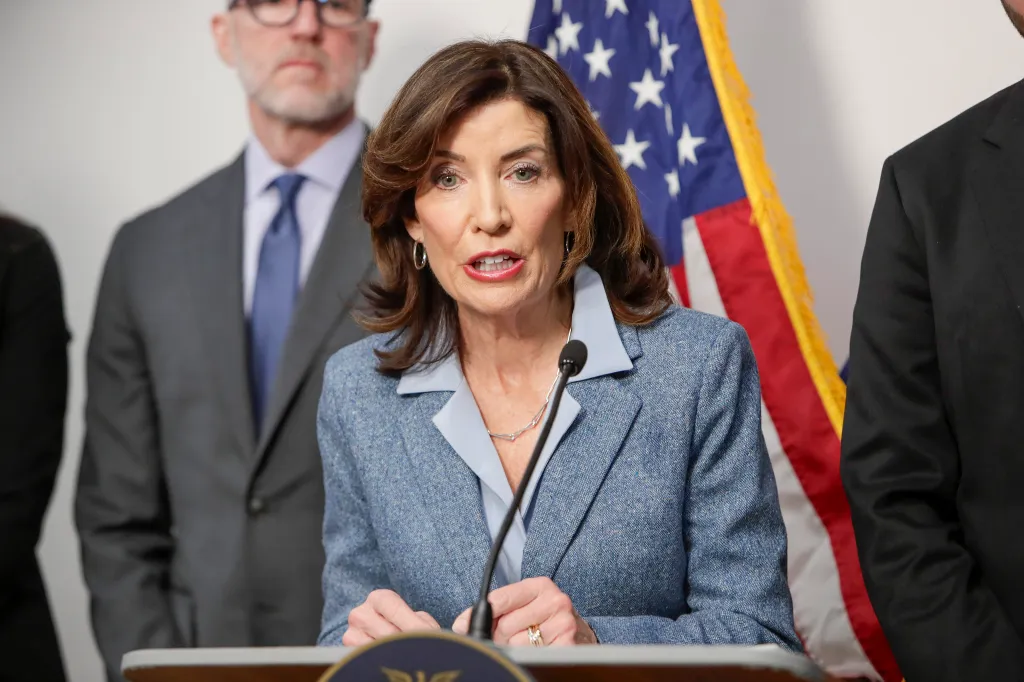 New York State Governor Kathy Hochul speaks at a podium with two men behind her, with an American flag to her right.