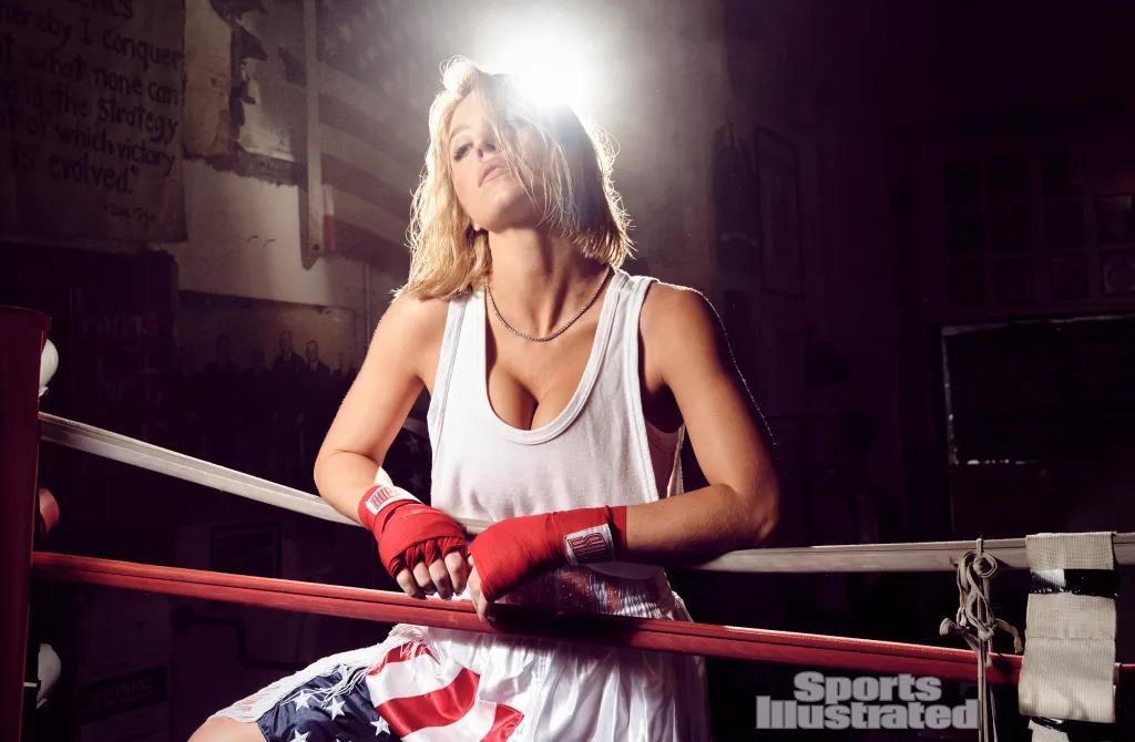 Sydney Sweeney leaning against the ropes of a boxing ring, wearing a white tank top, red boxing wraps, and boxing shorts with an American flag design.
