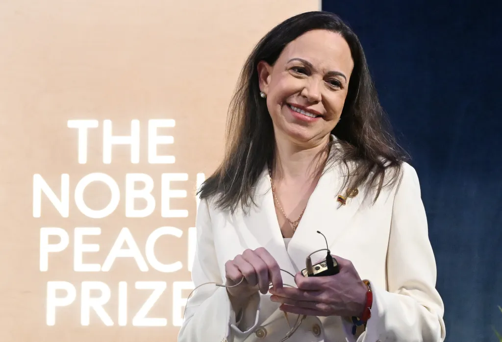 Maria Corina Machado, a Nobel Peace Prize recipient, smiles while holding eyeglasses and a microphone pack at a press conference.