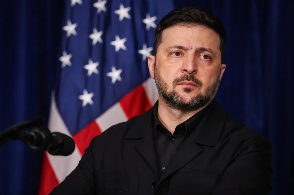 Ukrainian President Volodymyr Zelenskyy speaking at a press conference with an American flag in the background.