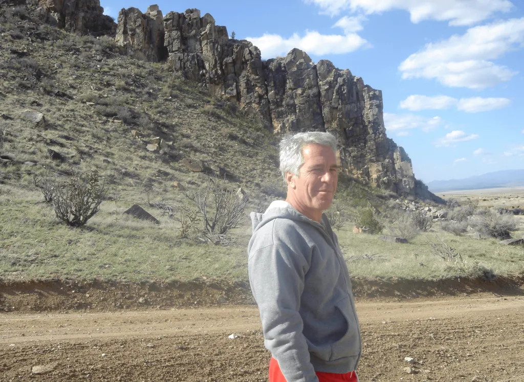 Jeffrey Epstein in a gray hoodie and red shorts stands on a dirt road in a desert landscape.