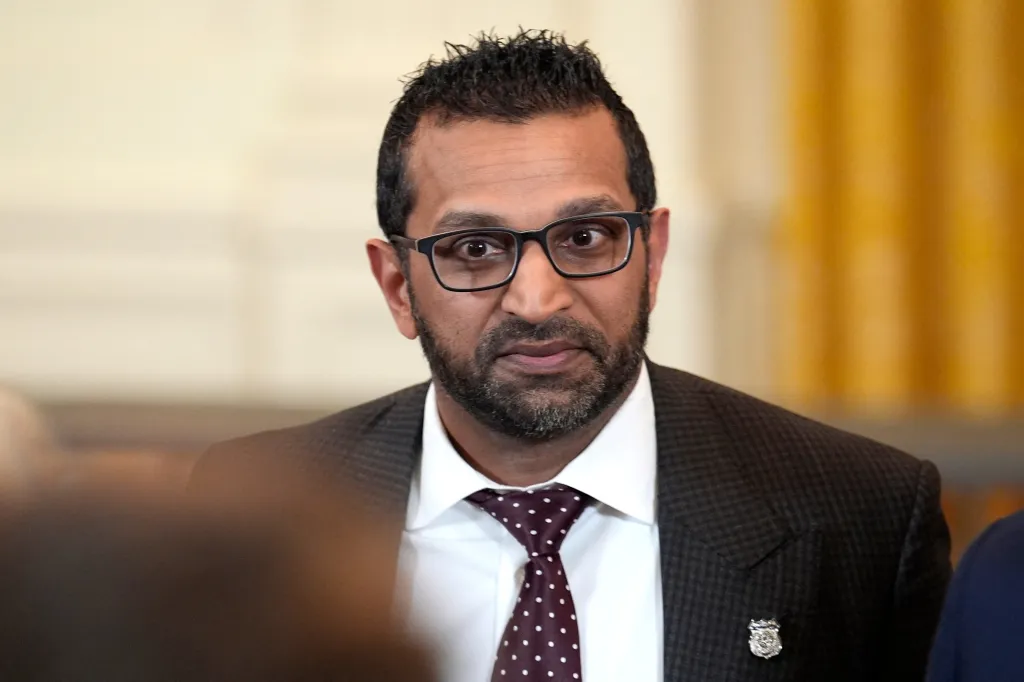 Kash Patel wearing glasses and a suit with a spotted tie and a small badge.