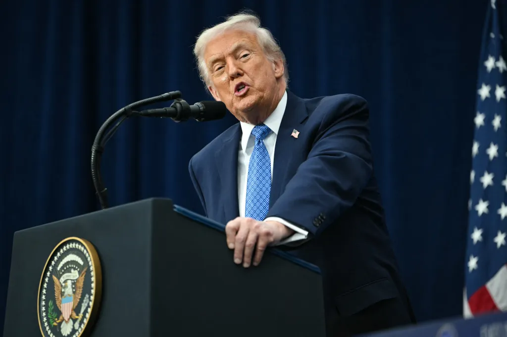 US President Donald Trump speaking at a podium with a microphone, with a US flag in the background.