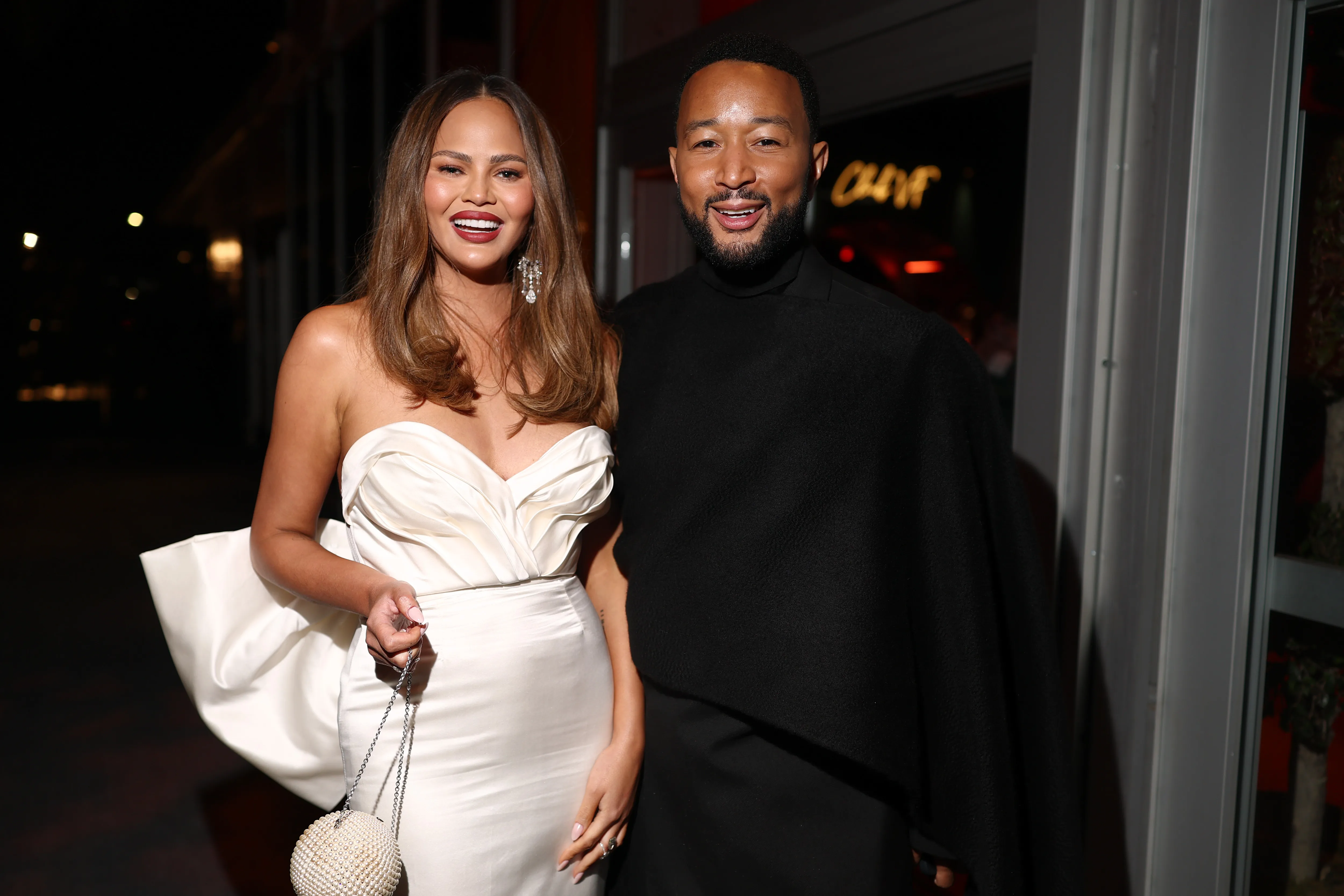 Chrissy Teigen in a cream strapless dress and John Legend in a black outfit smiling at the 2025 Vanity Fair Oscar Party.