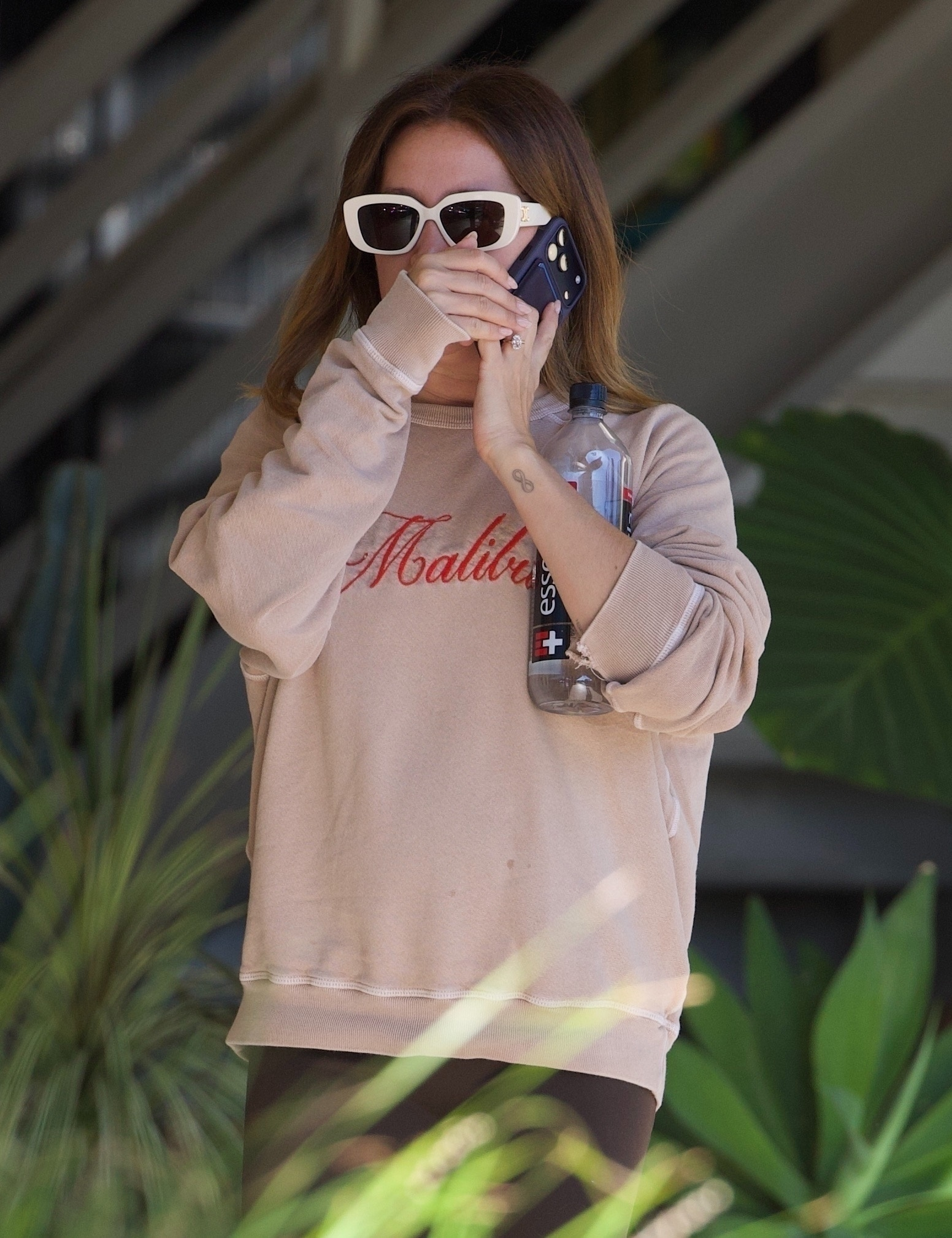 Ashley Tisdale on the phone wearing sunglasses and a Malibu sweatshirt, holding a water bottle.