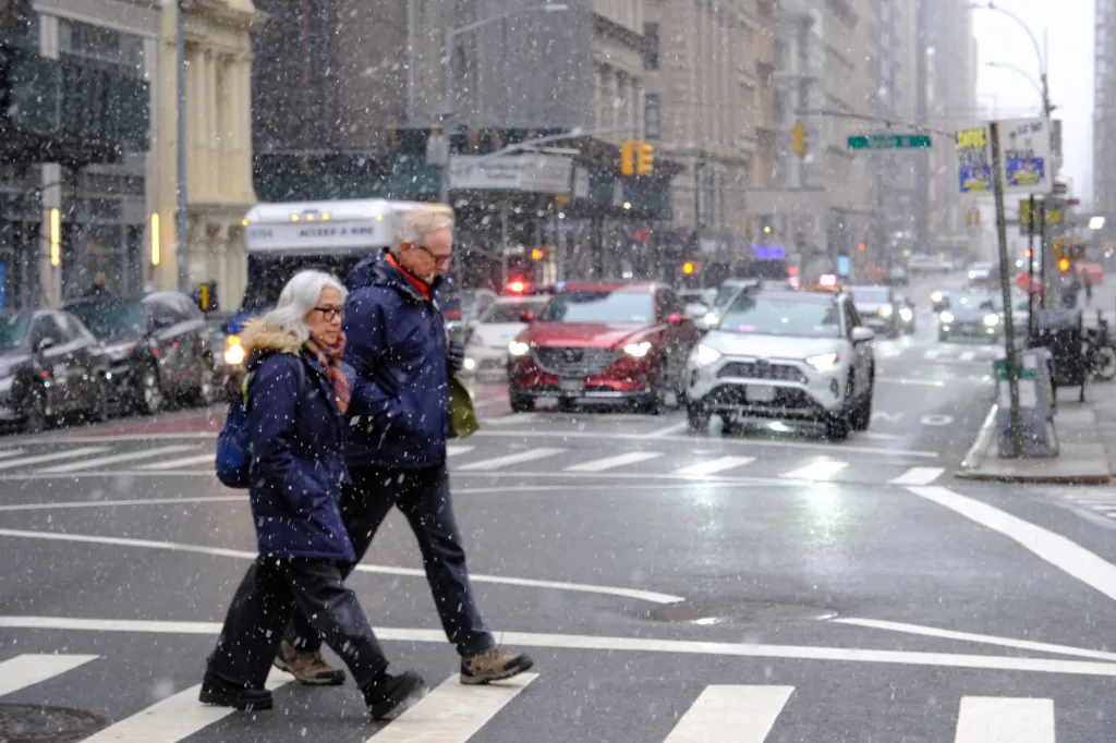 Two pedestrians, bundled in winter coats, cross a snowy New York City street with cars and buildings in the background.