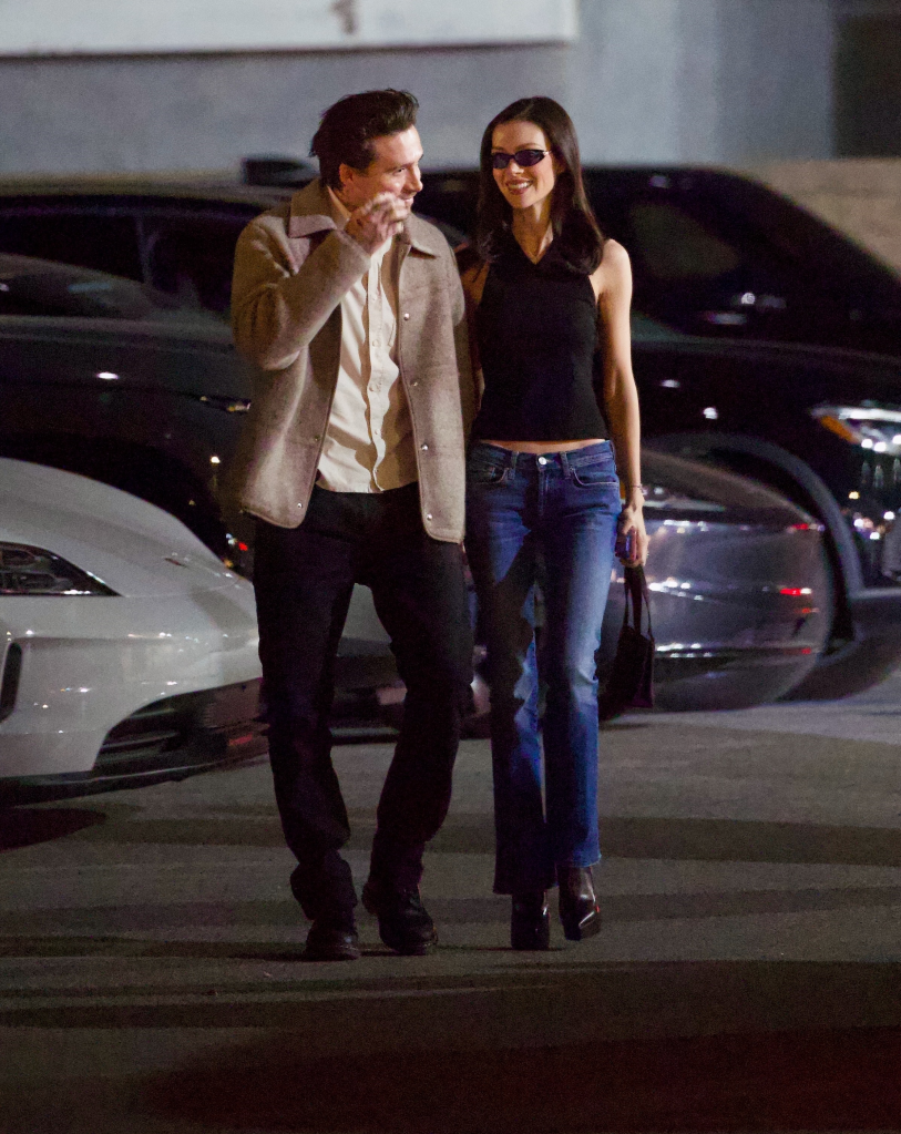 Brooklyn Beckham in a tan jacket and black pants, and Nicola Peltz in a black sleeveless top and blue jeans, smiling and holding hands while walking outside at night.
