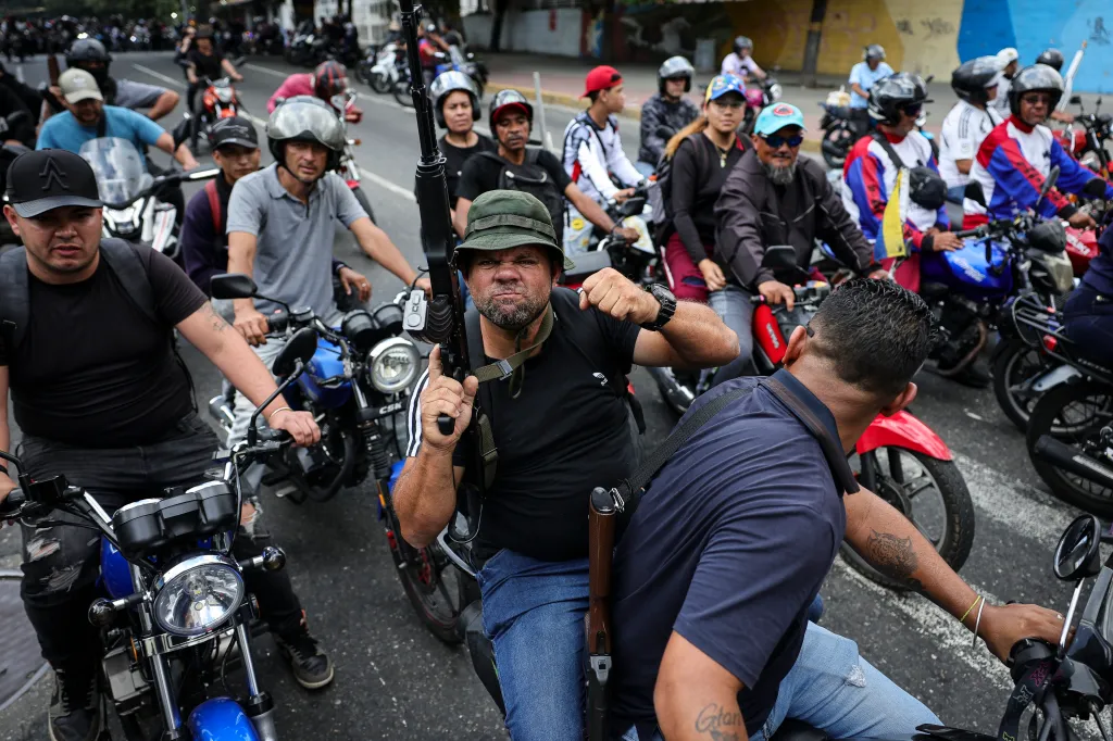 A man holding an assault rifle and clenching his fist, riding a motorcycle in a caravan of armed men on motorcycles.