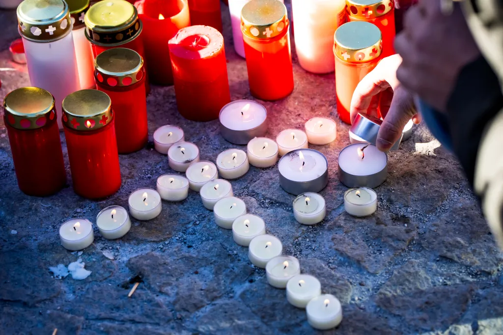A hand lights candles arranged in a heart shape on a stone ground, surrounded by taller red and white candles in glass holders.