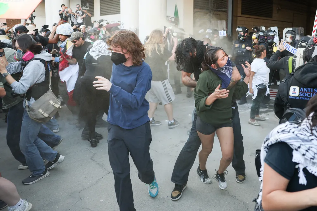 Protesters, some wearing respirators, flee a cloud of gas while a man protects a woman in an immigration protest.