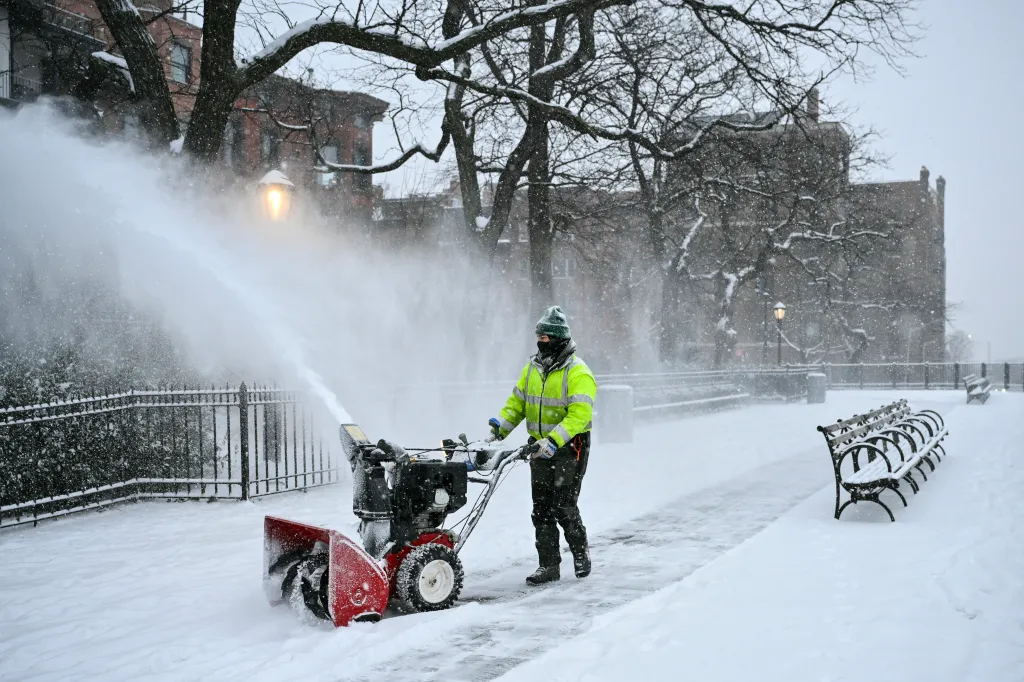 A worker in a bright yellow jacket uses a snowblower to clear a snow-covered path.