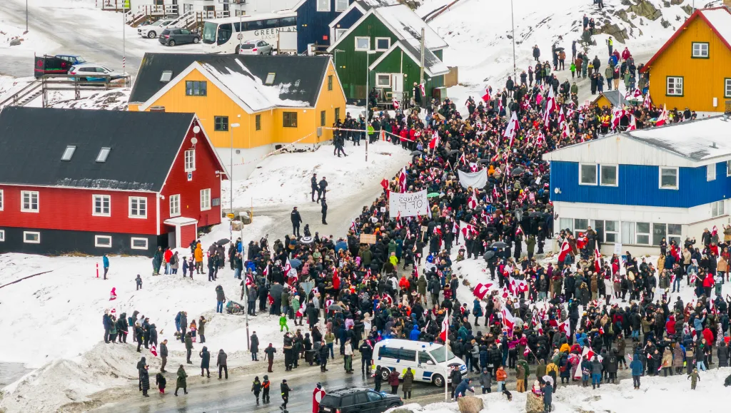 Aerial view of a large crowd of people protesting, many waving Greenlandic flags, on a snowy street in Nuuk, Greenland, with colorful houses visible on the hillside.