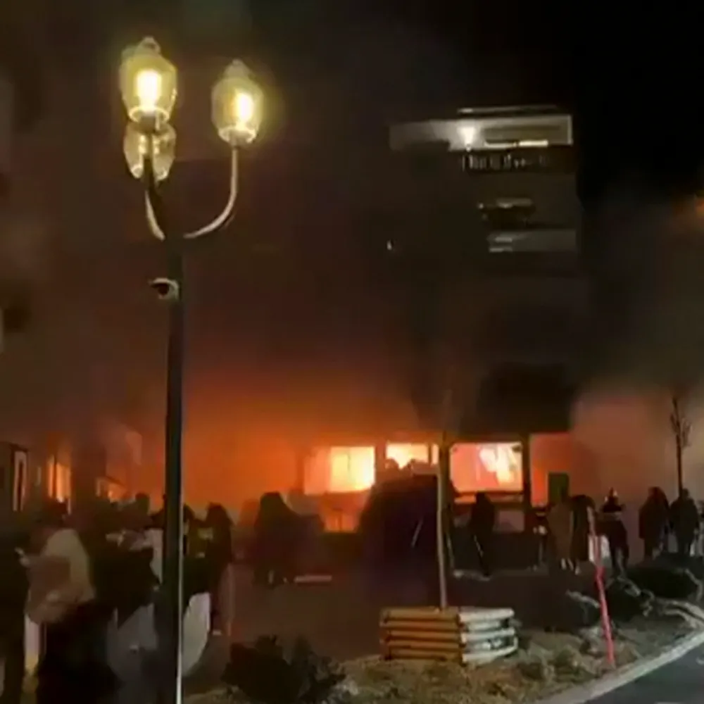 Building on fire at night, with bright orange flames visible through windows and smoke rising into the dark sky, people watching nearby.