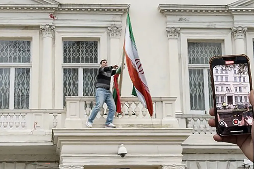 A protester pulls down an Iranian flag from the balcony of the Iranian Embassy in London while a person records on a smartphone.