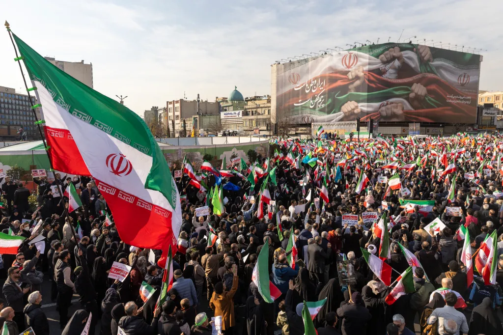 Thousands of pro-government Iranian demonstrators wave Iranian flags in Tehran's Enqelab Square, with a large banner depicting two hands clenching a flag in the background.