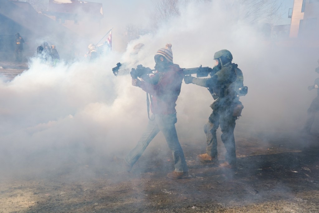 Two federal officers in gas masks and tactical gear hold rifles amid tear gas.