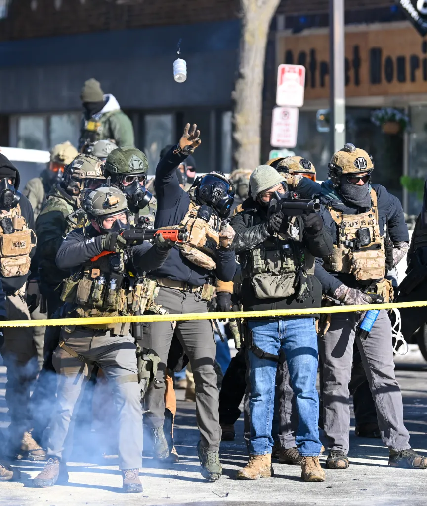 Federal officers in tactical gear and gas masks confront protestors, one throwing a tear gas canister while another fires less-lethal rounds.