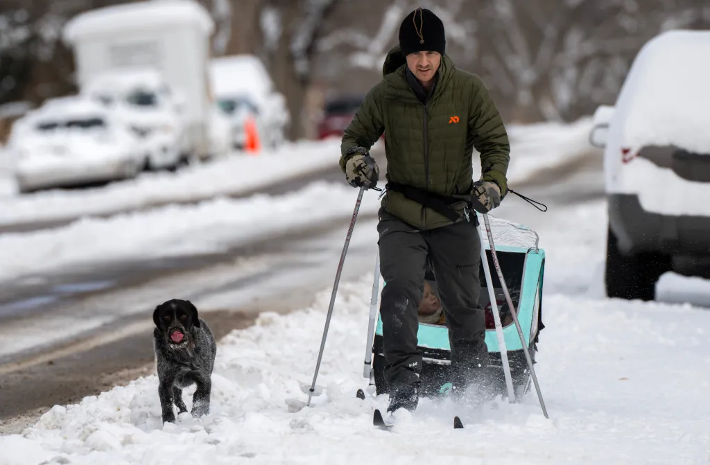 Man on skis pulling a trailer with children in the snow, with a dog running alongside.