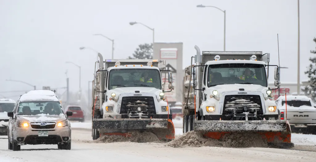 Two snow plows clear snow from a highway as a car drives by.