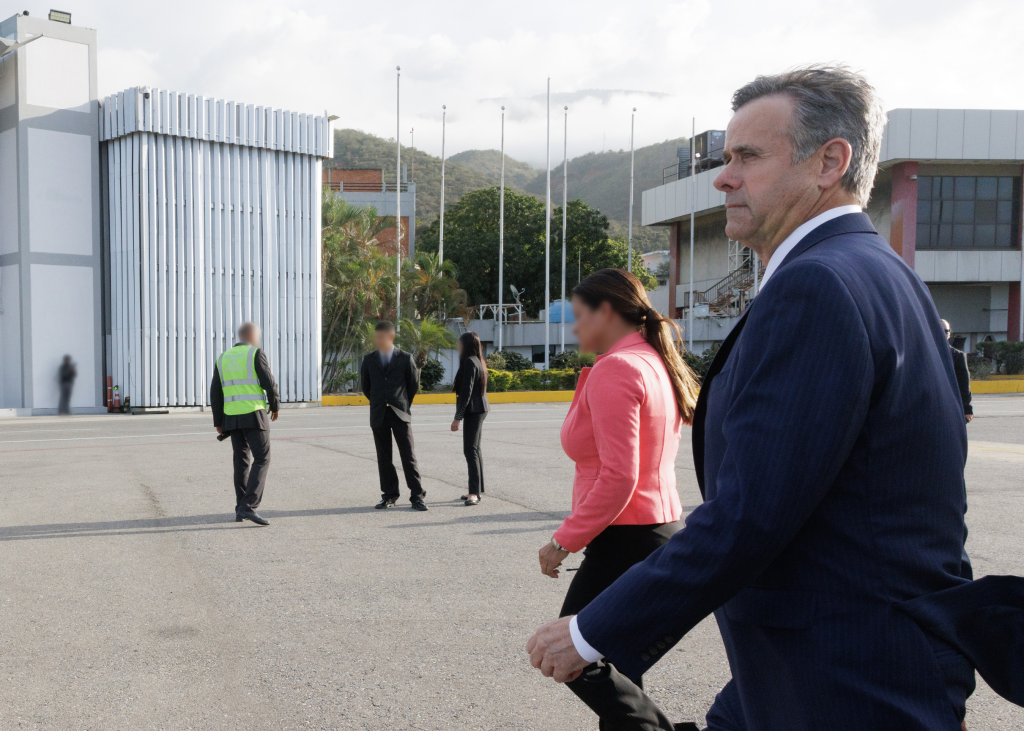 A man in a blue suit walks past other people on an asphalt ground with buildings and hills in the background.