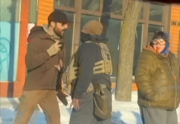 Three men in winter attire, one wearing tactical gear, stand outside a building in the snow.