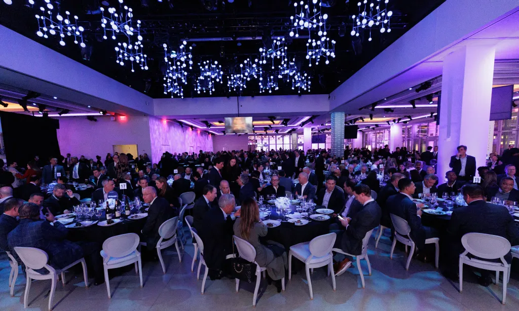 Attendees gather at tables in a large event space with blue lighting and modern chandeliers.