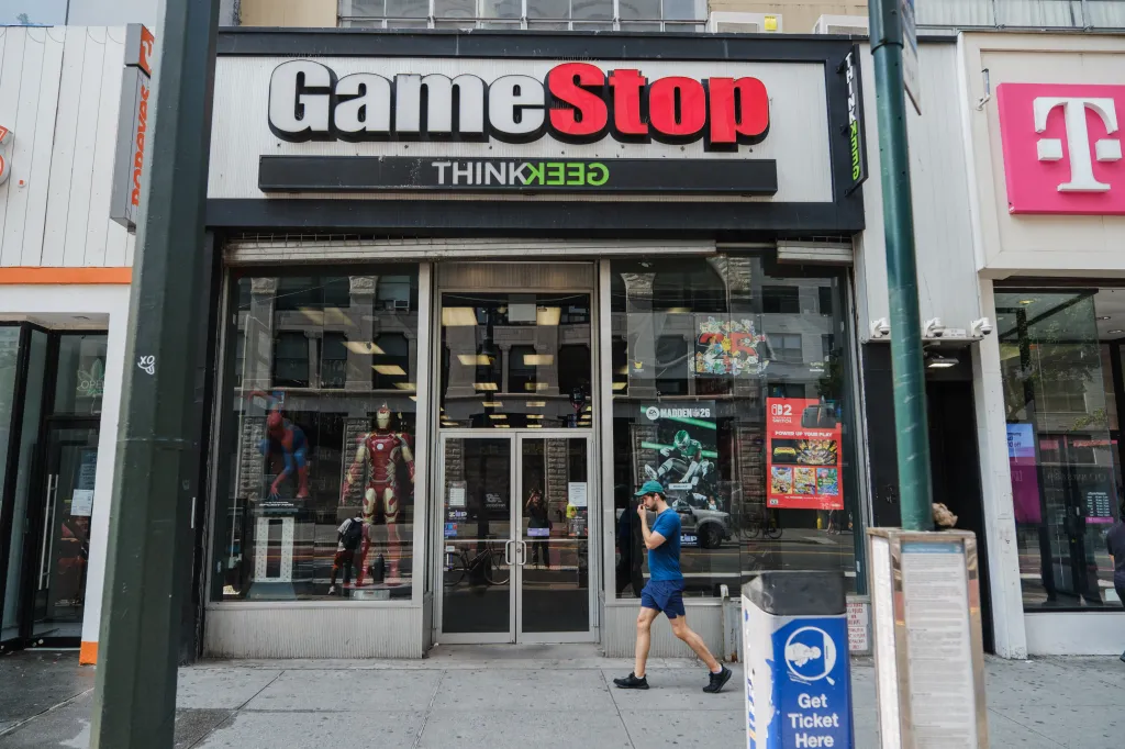 A GameStop store with displays of Spider-Man, Iron Man, and various Nintendo Switch games in the window, and a man walking by on the sidewalk.