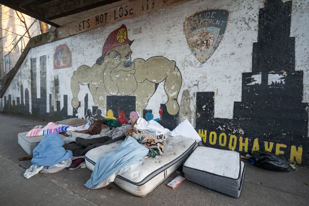 Homeless encampment with mattresses and blankets under a bridge, with graffiti of a bulldog and 