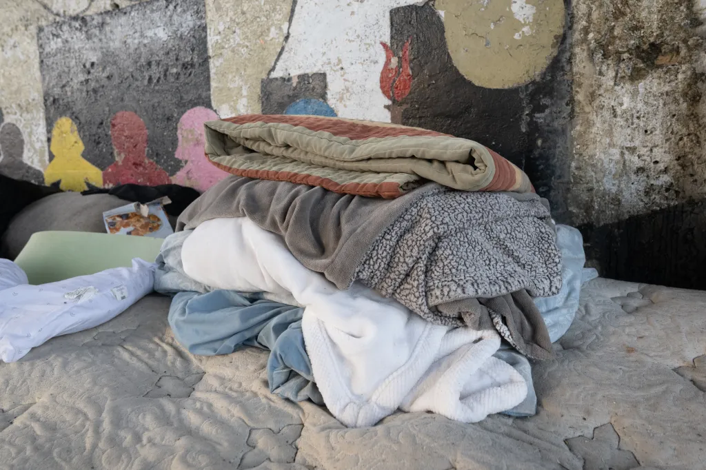 Folded linens on a mattress in a homeless encampment under a train bridge.