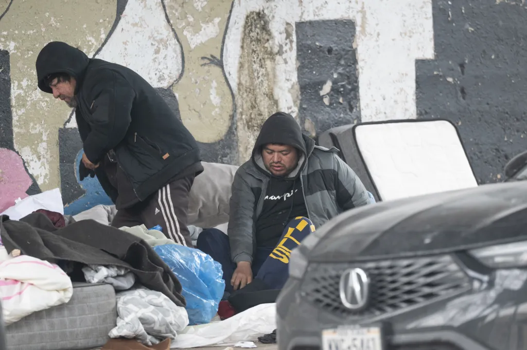 Two unhoused men in hooded jackets sit amidst their belongings, including mattresses and blankets, under a train bridge in Queens, NY.