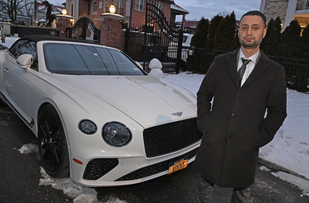 Moshe Borukh standing next to his white Bentley convertible on a snowy street.