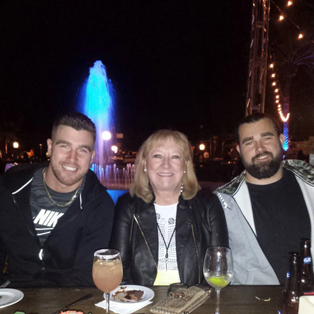 A woman and two men sit at a table outdoors at night with a blue fountain in the background.