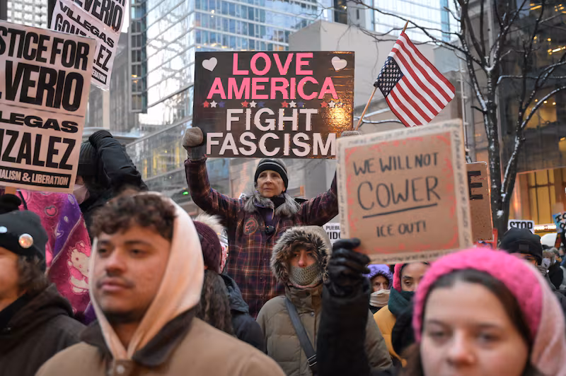 A woman stands elevated in the centre of a crowd holding a sign that says "LOVE AMERICA, FIGHT FASCISM"